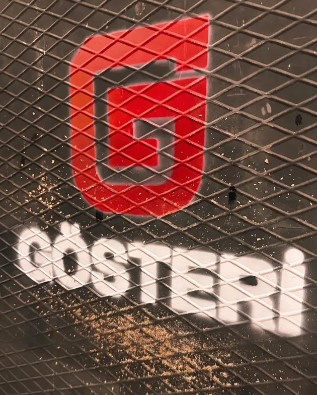 Gösteri Woodworks logosu, sac yüzey üzerine stencil ve sprey boya tekniğiyle uygulanmış; üzerinde atölyeden ahşap tozu detayı.