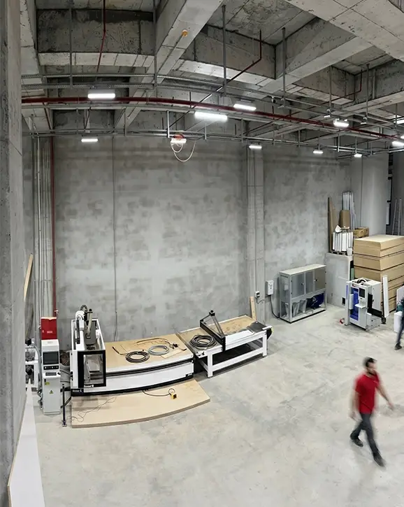Gösteri Woodworks atölyesinde CNC işleme merkezi ve eğri formlu özel kesim ahşap parça; sağda hareket halinde operatör.