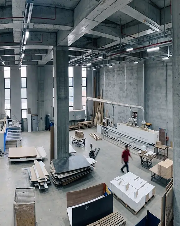 Gösteri Woodworks üretim tesisinde kenar bantlama hattı ve istiflenmiş ahşap paneller; arka planda hareket halindeki operatör.